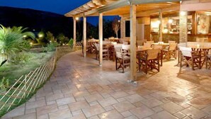 Un patio al aire libre con mesas y sillas de madera, una zona de estar cubierta y un suelo de baldosas de piedra.