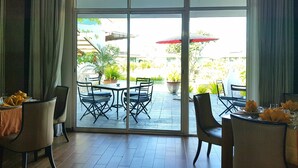 Un comedor con mesas y sillas, un amplio ventanal corredero que da a un patio exterior con sombrillas y plantas en macetas.