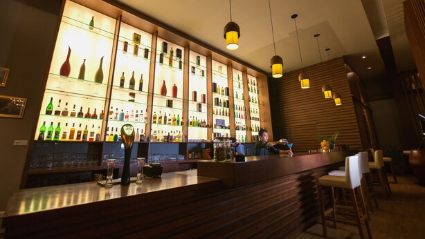 Un bar con estantes llenos de botellas y vasos, barra de madera y lámparas colgantes.