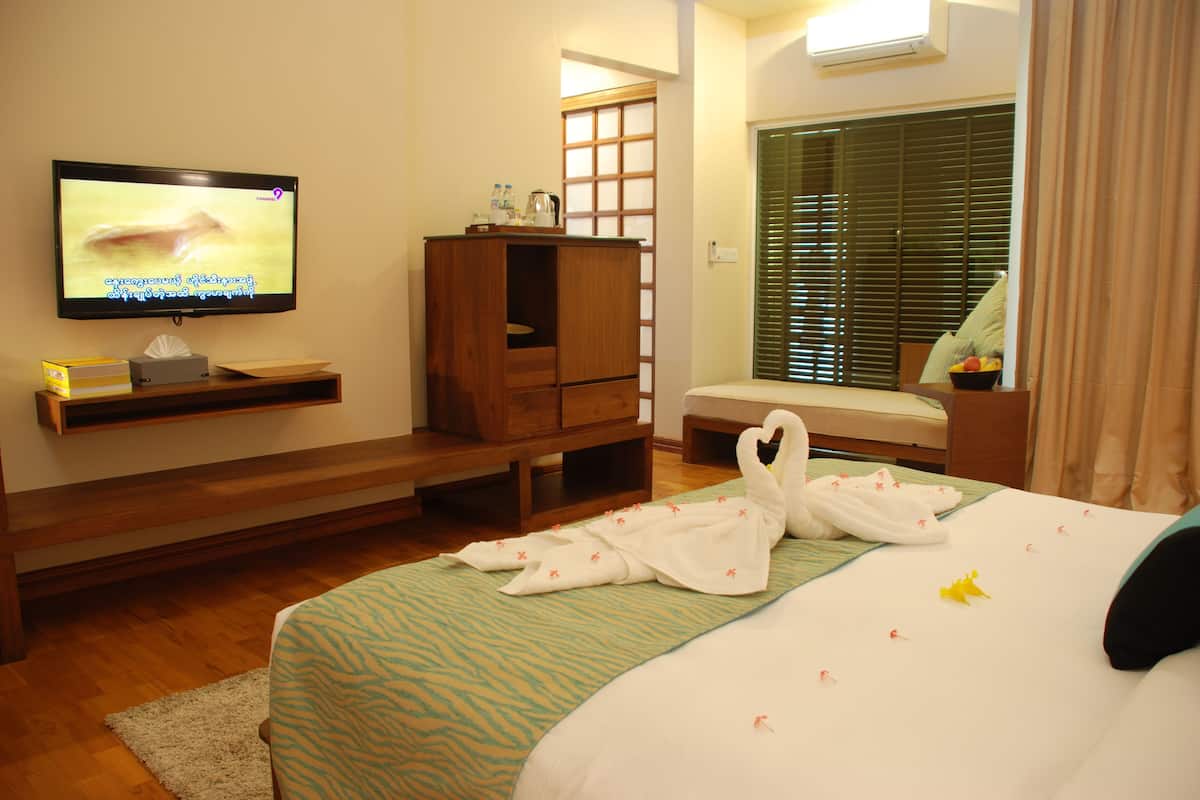 Habitación de hotel con televisión en la pared, un armario de madera, una cama con almohada con forma de cisne y una ventana con persianas.