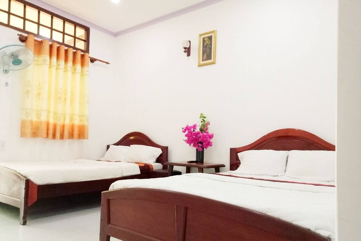 A hotel room with two wooden beds, a small table, a wall-mounted fan, and a window with a curtain.