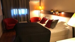 Une chambre d’hôtel avec un lit, deux chaises rouges, une lampe et une fenêtre avec des rideaux.
