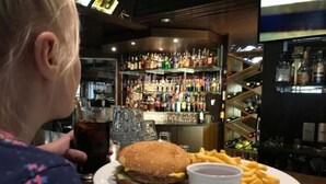 Une personne assise au bar, devant une assiette de nourriture comprenant un hamburger, des frites et une boisson.