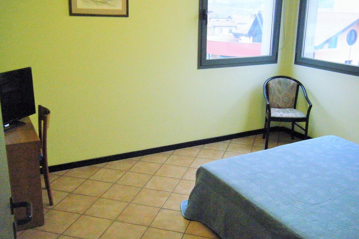Une pièce comprenant un lit, une chaise, un bureau avec un ordinateur, une fenêtre et un tableau encadré accroché au mur.
