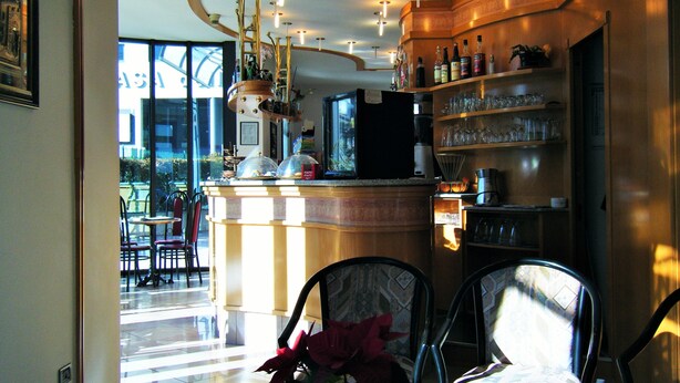 A modern bar area with a curved ceiling, a refrigerator, and shelves with glasses and bottles. There are chairs and a table in the foreground, and a view of the outside through large windows. - Residence Vezzoli (Darfo Boario Terme)