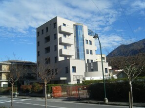 Un bâtiment moderne à plusieurs étages, doté d’une tour cylindrique en verre, entouré d’arbres et d’une clôture.