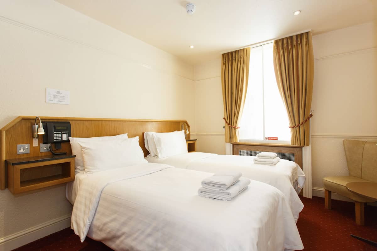 A hotel room with two beds, a wooden headboard, a bedside telephone, a window with curtains, and a chair.