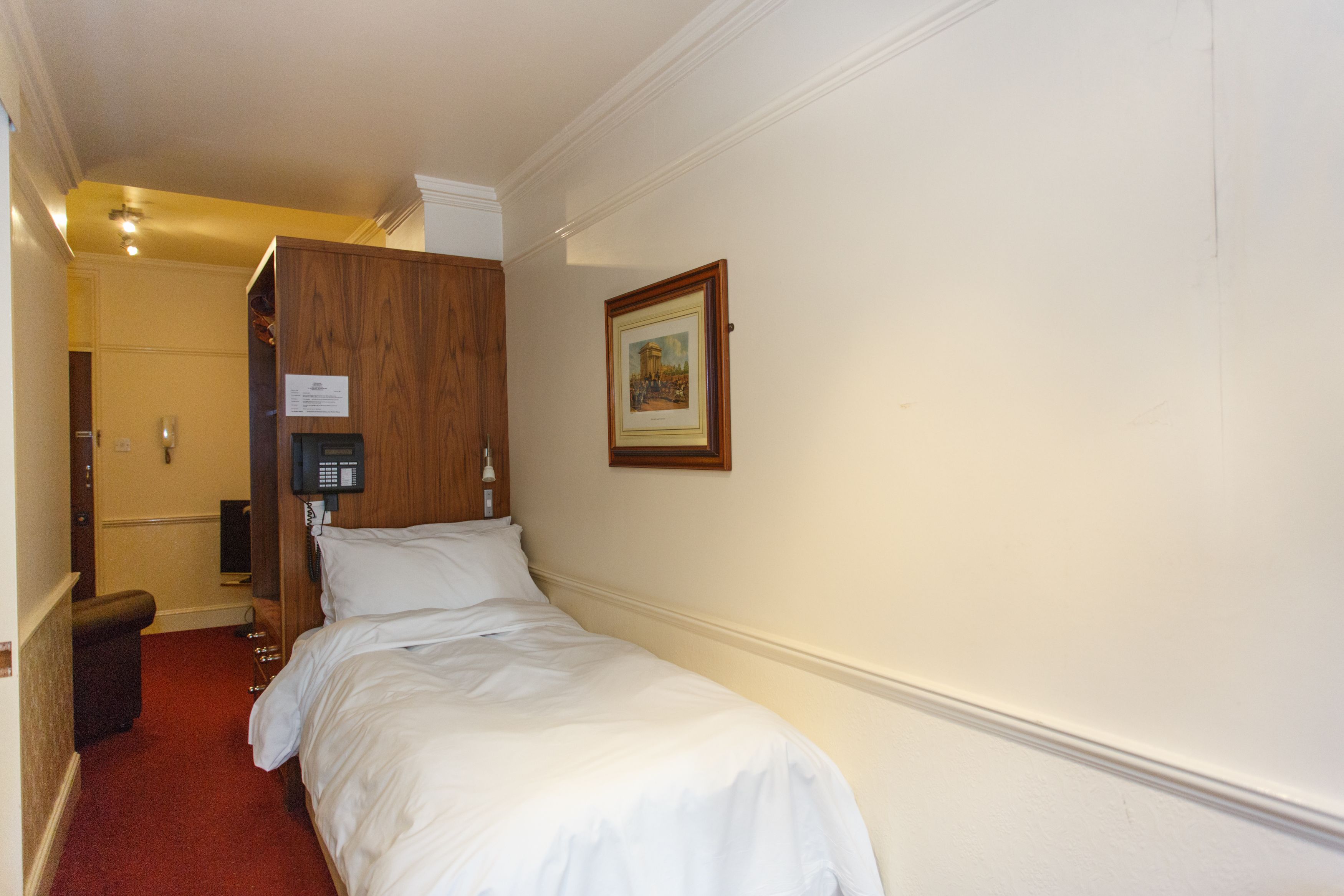 A hotel room with a single bed, a wooden wardrobe, a framed picture on the wall, and a telephone on the wall.