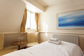 Una habitación de hotel con cama, una mesita redonda con dos vasos, una silla y una ventana con cortinas.