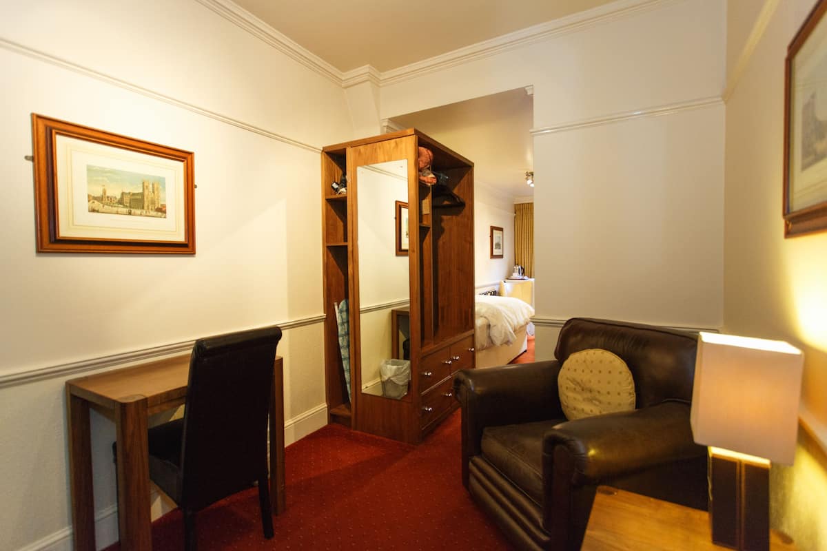 A hotel room with a wooden wardrobe, a desk, a leather armchair, and a bedside table with a lamp.