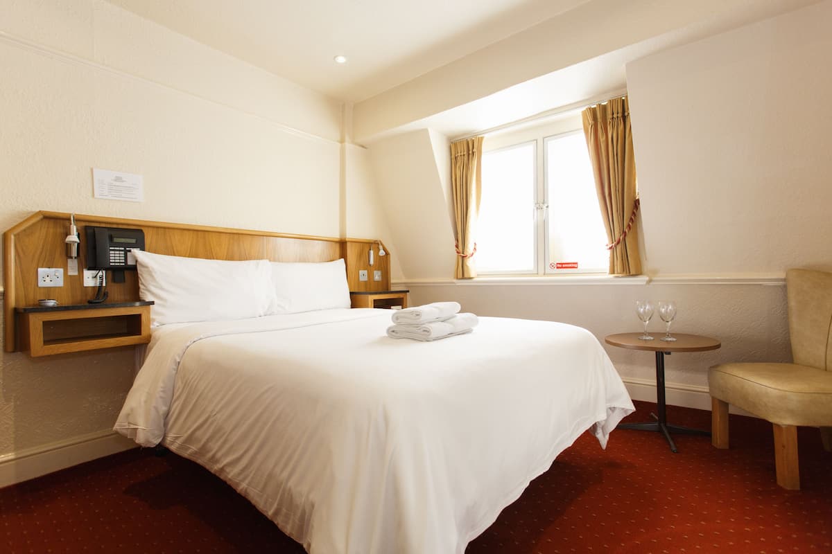 A hotel room with a bed, a bedside table with a phone and a lamp, a small round table with two glasses, and a chair.