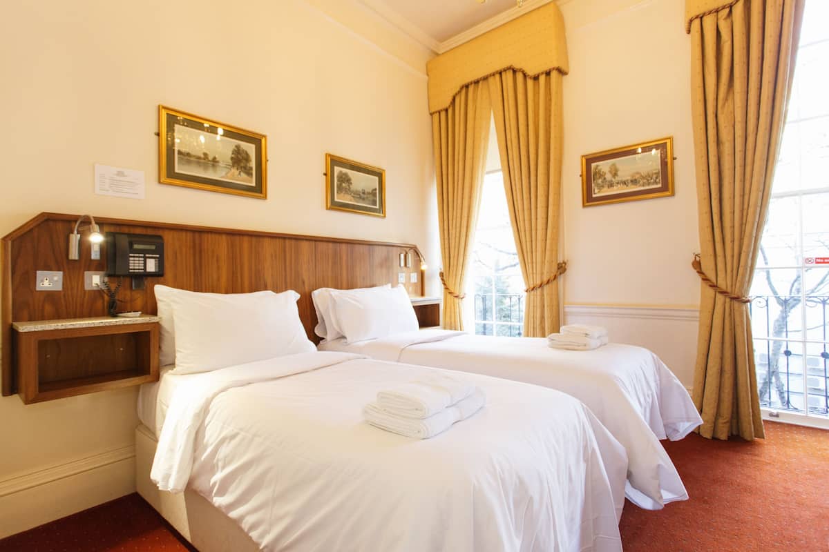 A hotel room with two beds, a wooden headboard, a bedside table with a lamp, and two framed pictures on the wall.