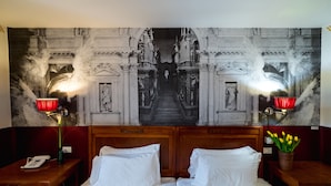 Een hotelkamer met een groot wandtapijt waarop een historisch tafereel afgebeeld is, twee nachtkastjes met lampen en een bed met witte lakens.