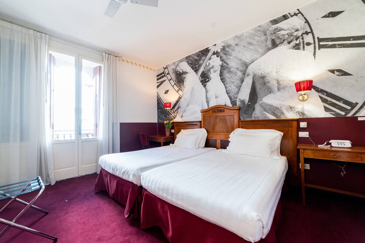 Een hotelkamer met twee bedden, een groot wandtapijt, een bureau met lamp en een raam met gordijnen.
