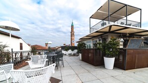 Een terras buiten met witte stoelen en een tafel, een houten toonbank met een prikbord, en een uitzicht op een klokkentoren in de verte.