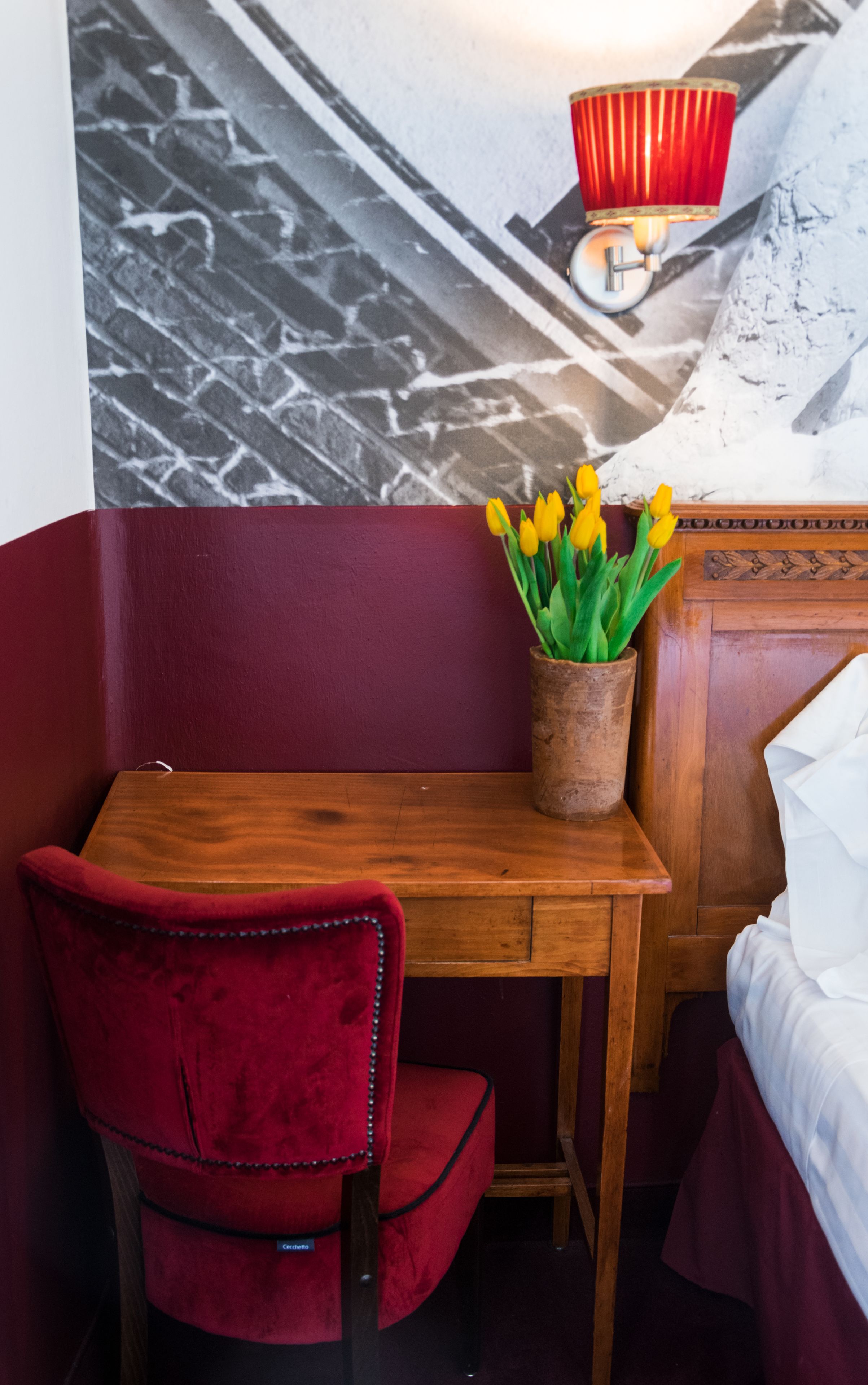 A room with a wooden desk, a red chair, a potted plant with yellow flowers, a red lamp, and a bed with white bedding.