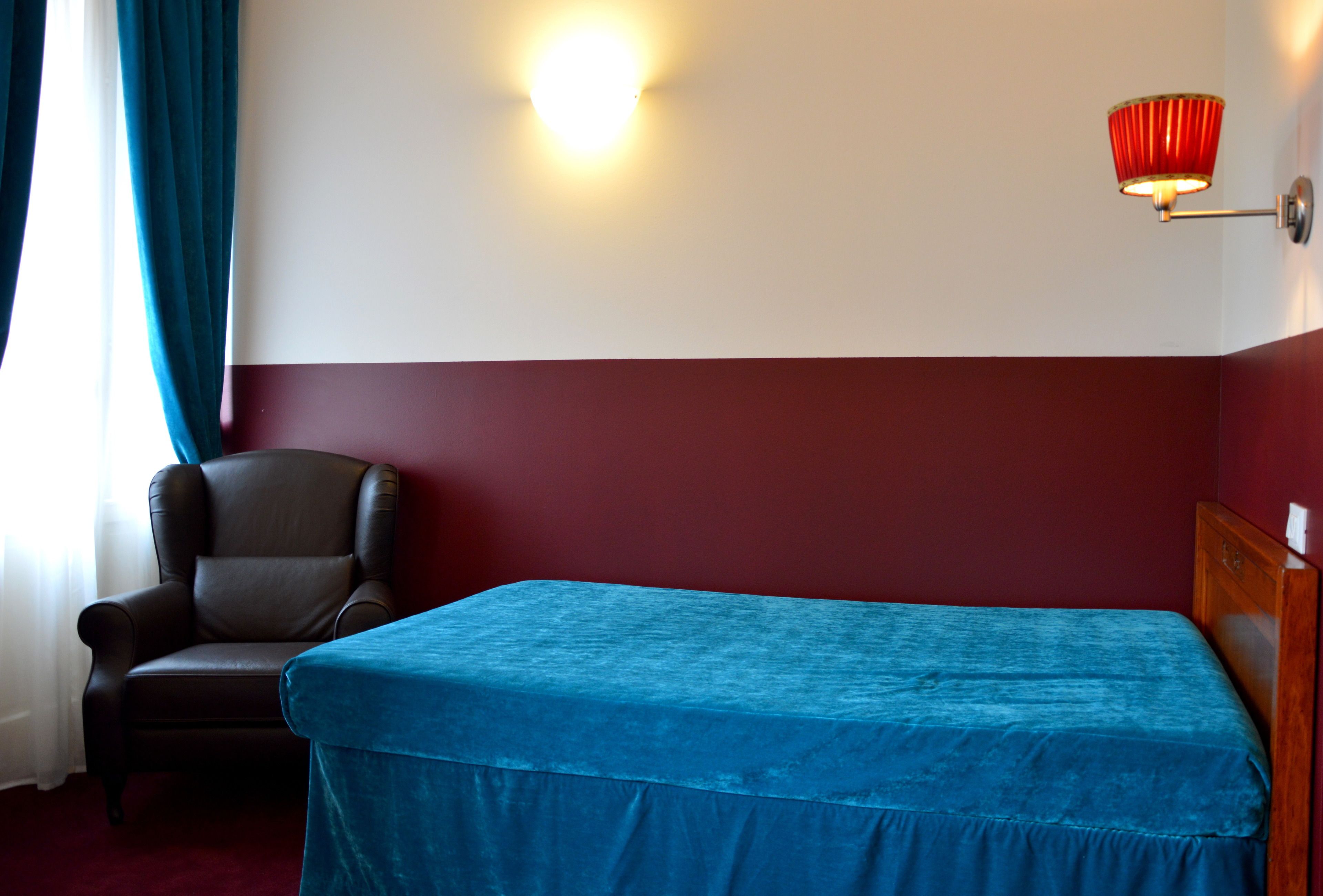 Una camera d'albergo con un letto, una sedia, una lampada a parete e una finestra con tende.