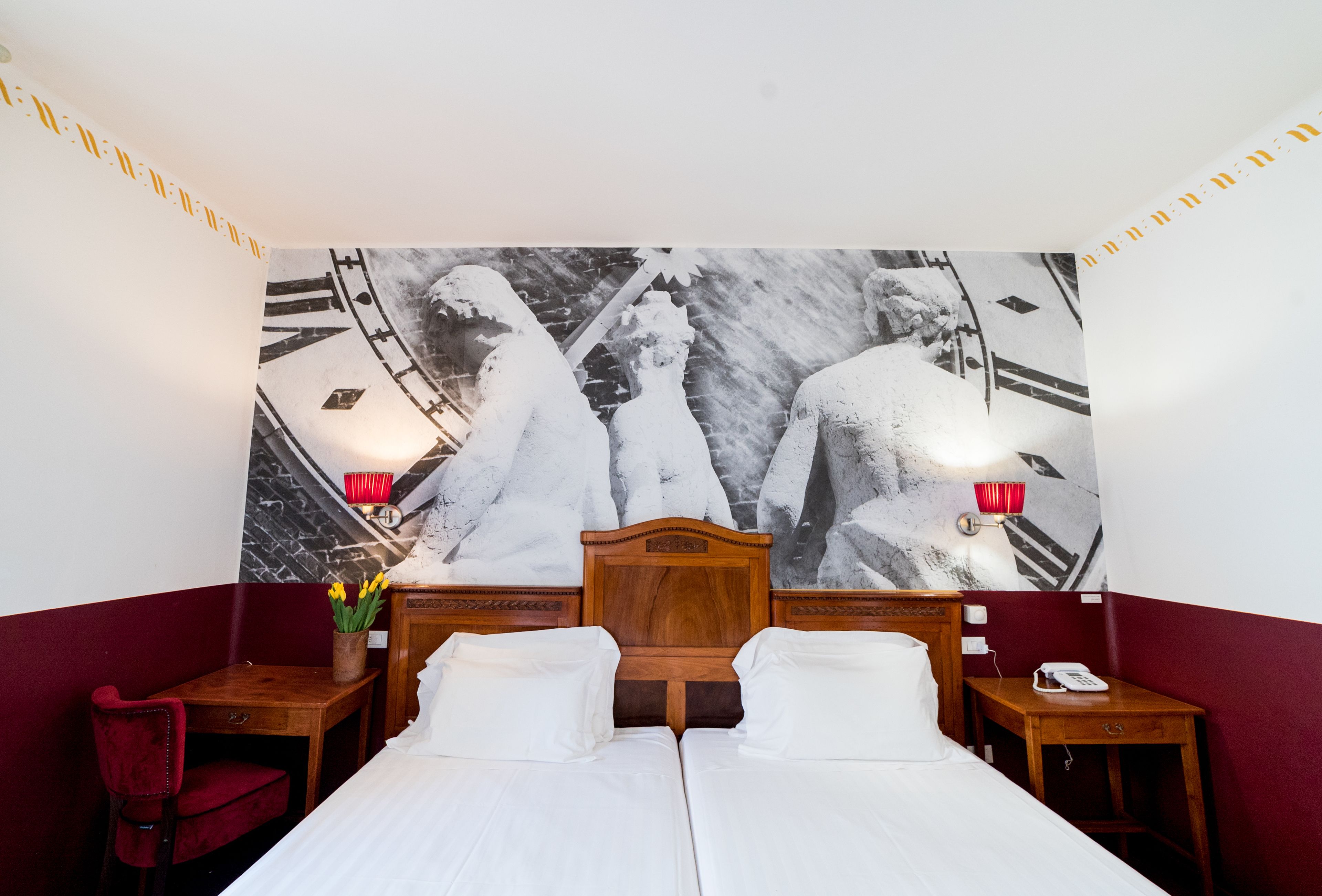 Une chambre d’hôtel avec un grand lit, deux tables de chevet, un fauteuil rouge, un vase avec des fleurs et une tapisserie murale.