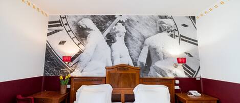 Habitación de hotel con una cama grande, dos mesitas de noche, una silla roja, un jarrón con flores y un tapiz en la pared.