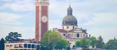 Um edifício histórico com uma torre de relógio e uma cúpula, rodeado por vegetação exuberante e um céu claro.