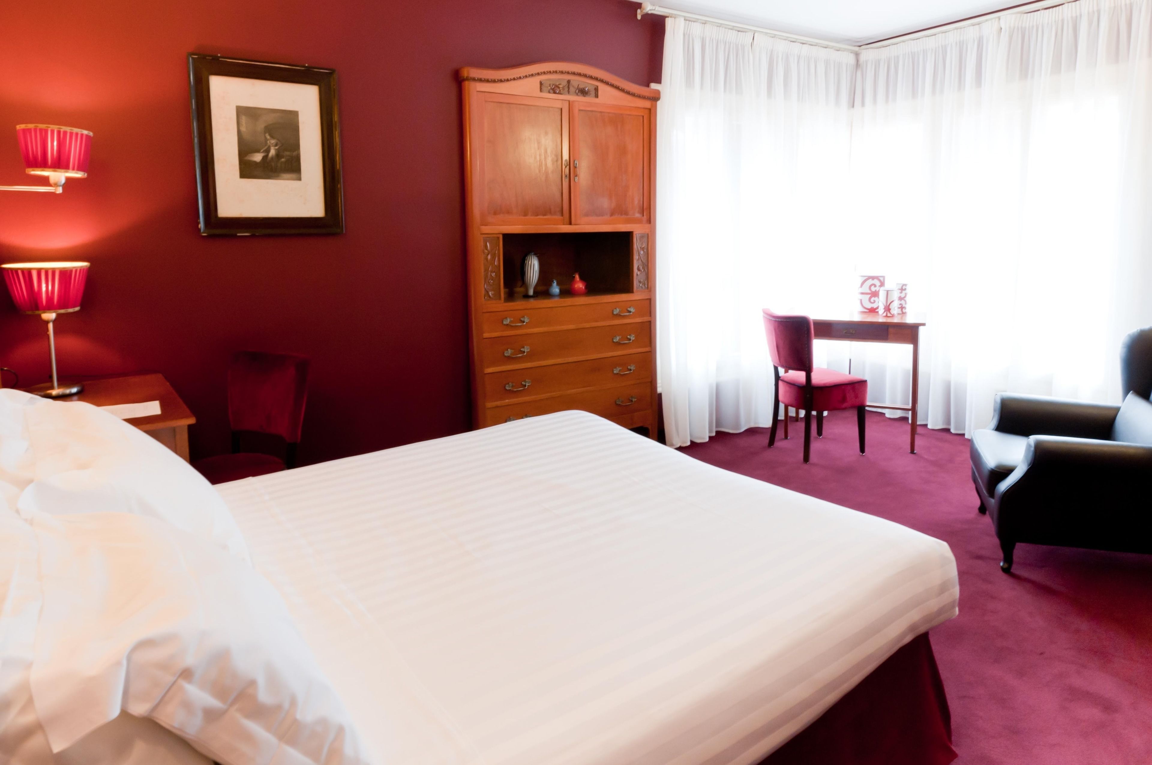 A hotel room with a large bed, a wooden wardrobe, a desk with a chair, and a red armchair.