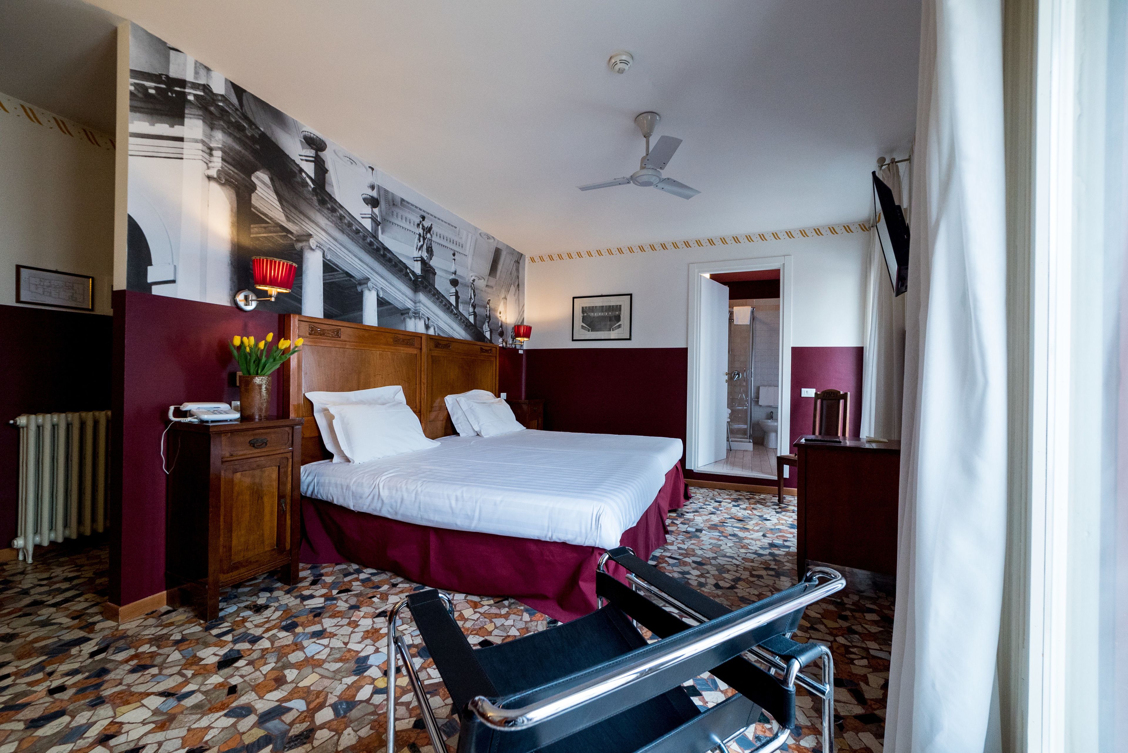 Ein Hotelzimmer mit einem großen Bett, einem Holfnachtkästchen, einem Schreibtisch, einem Stuhl, einem Heizkörper, einem Deckenventilator und einem Mosaikboden.
