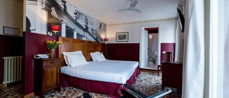 Una habitación de hotel con una cama grande, una mesita de noche de madera, un escritorio, una silla, un radiador, un ventilador de techo y un suelo de mosaico.
