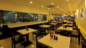 A restaurant with multiple tables and chairs, a counter with a display case, and a kitchen area visible through a glass partition.