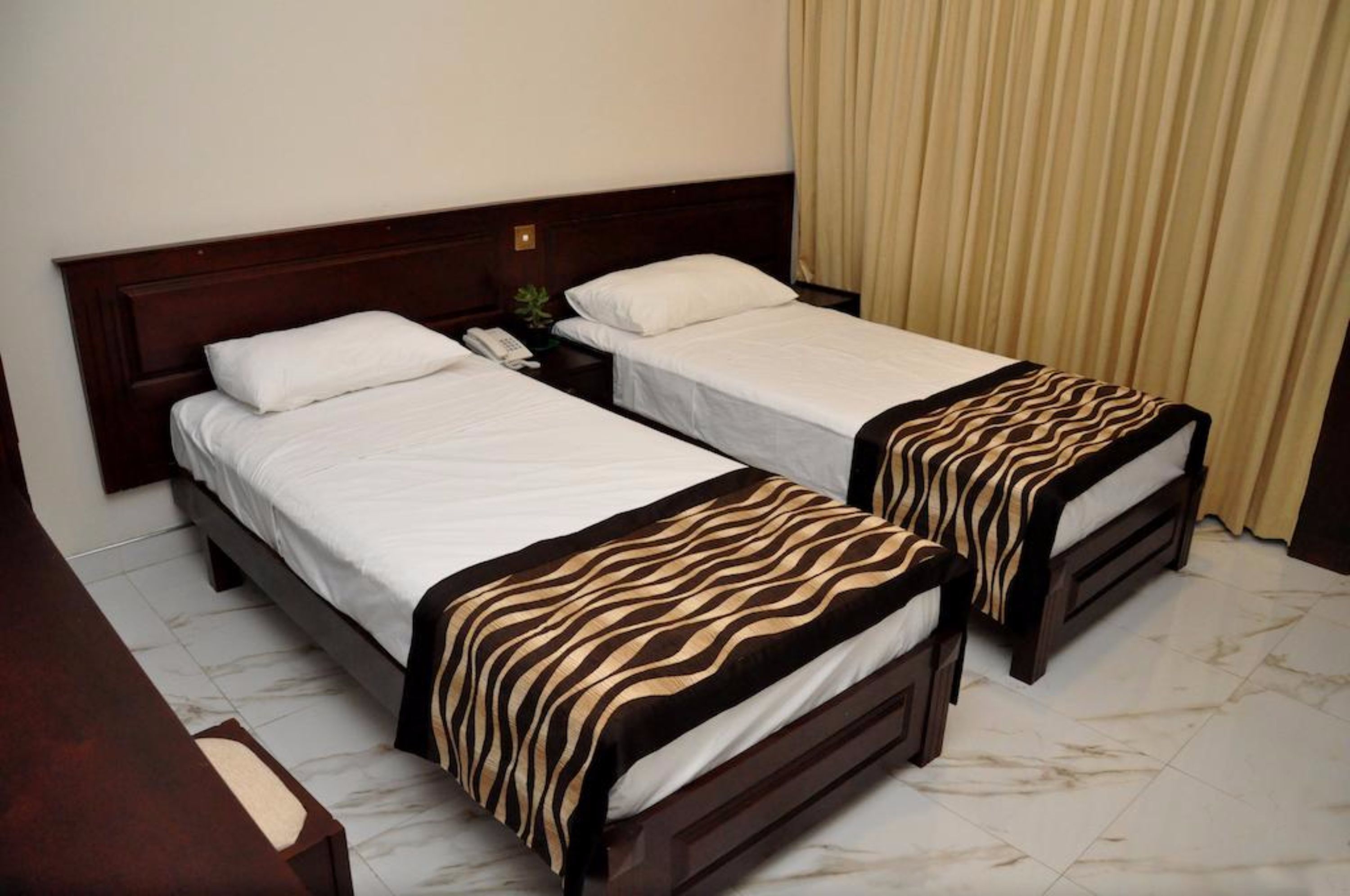 Habitación de hotel con dos camas individuales, cabecera de madera, mesita de noche con lámpara y cortina.