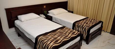 Habitación de hotel con dos camas individuales, cabecera de madera, mesita de noche con lámpara y cortina.