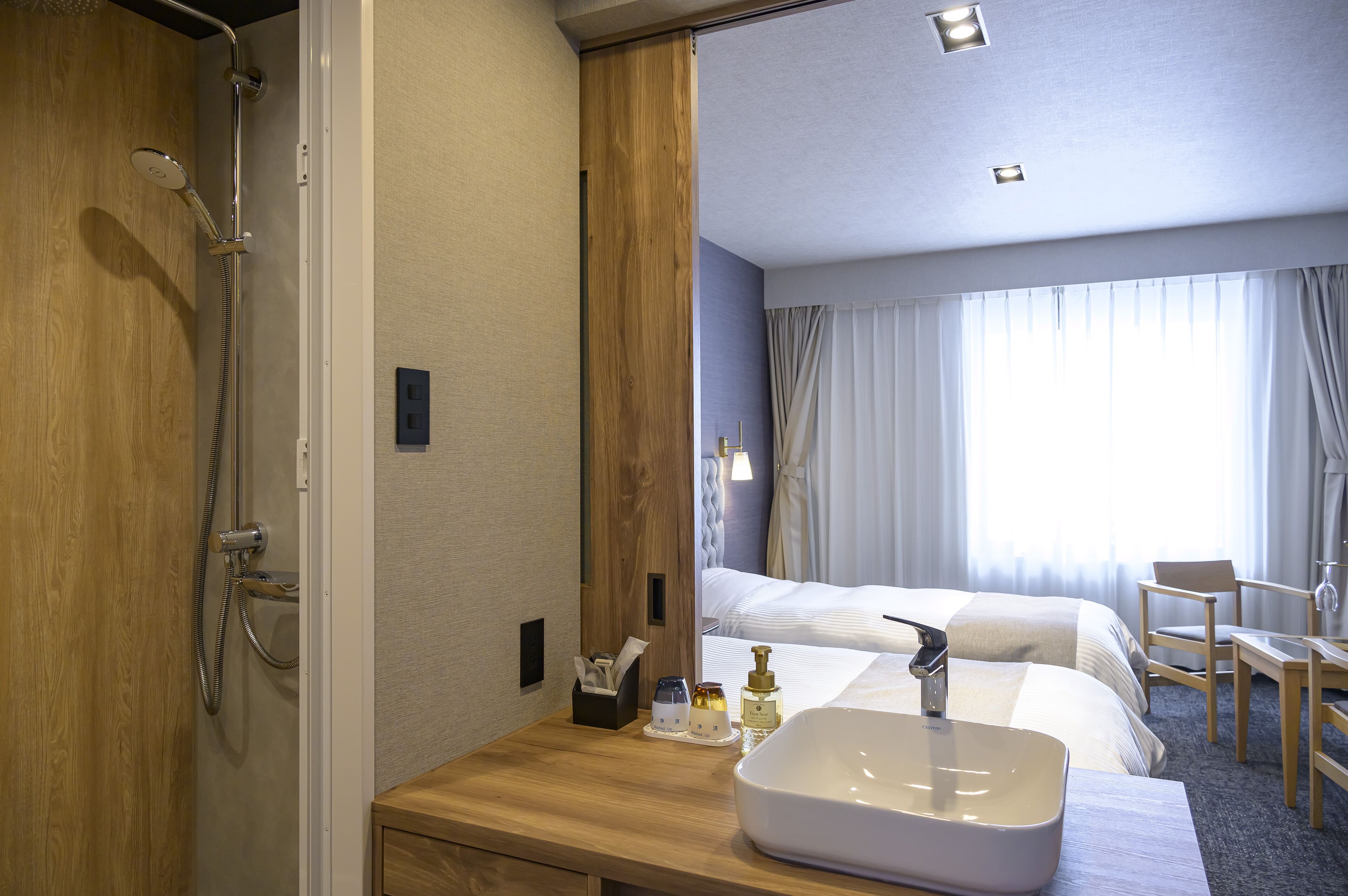 A hotel room with a wooden shower enclosure, a large bed with white linens, a bedside table with a white sink, and a view of a window with curtains.