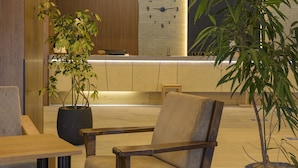 Zona de recepción con un mostrador de madera, una planta grande en maceta, un reloj y un área de descanso con una silla acolchada.