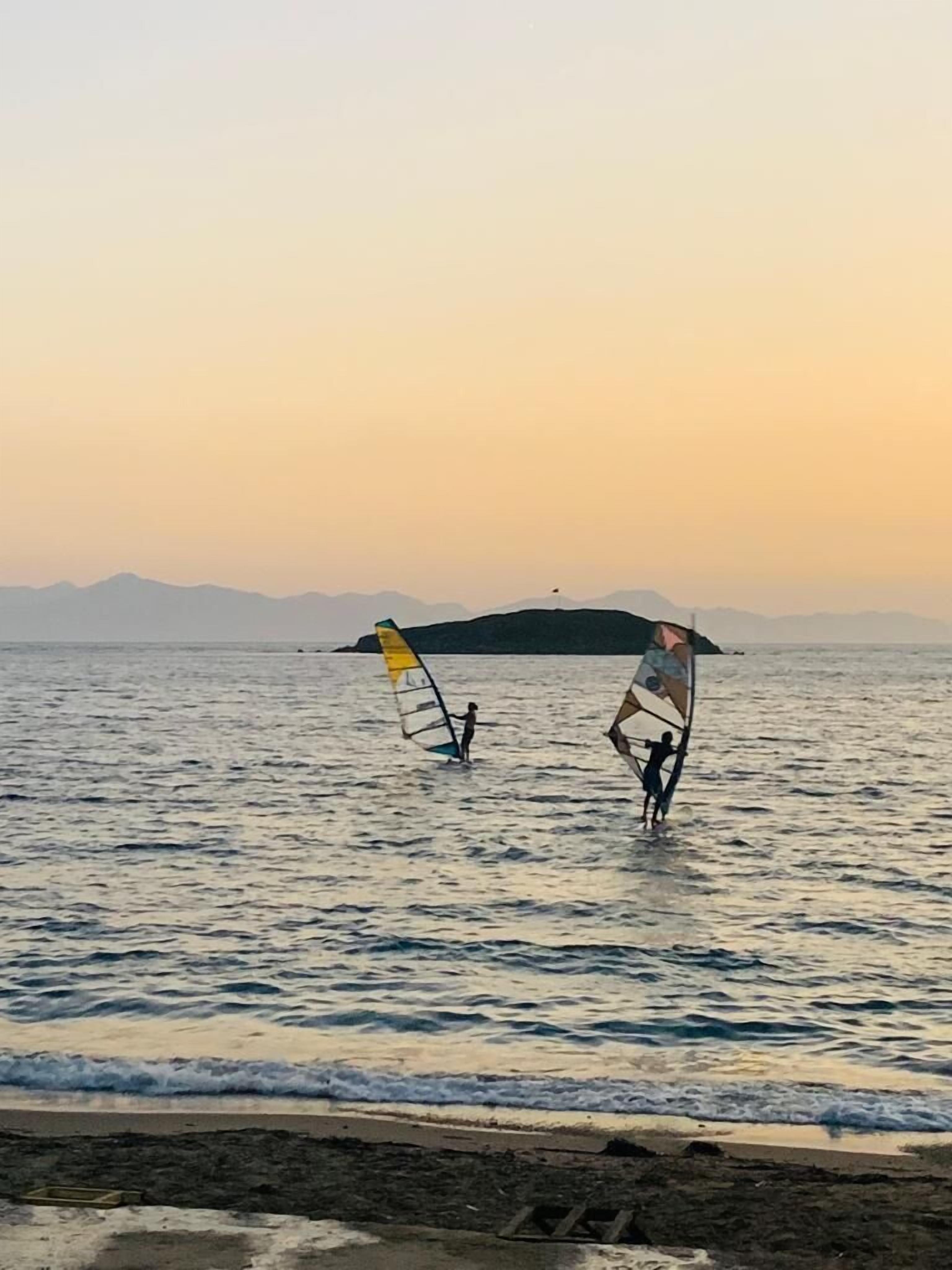 A beach scene with two windsurfing individuals and a sailboat in the background.