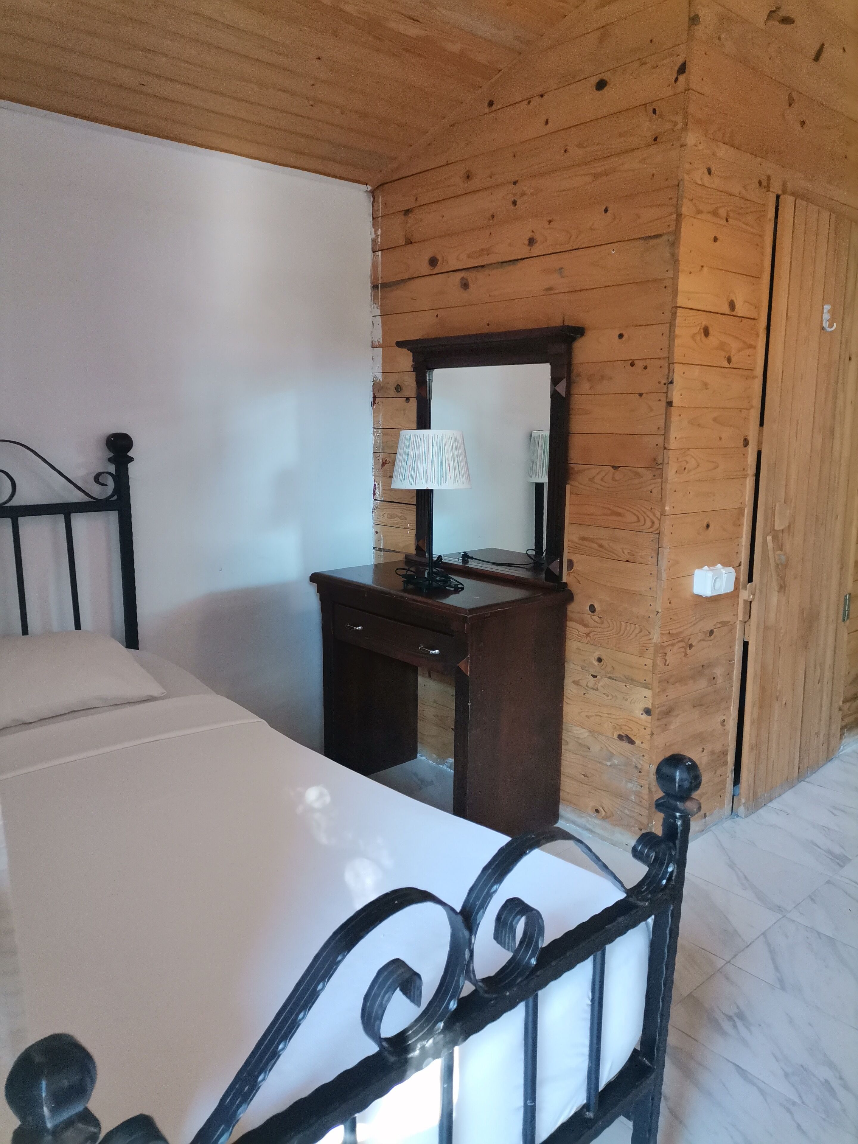Een slaapkamer met een houten plafond, een bed met een zwart hoofdeinde, een nachtkastje van hout met een lamp en een houten garderobe.