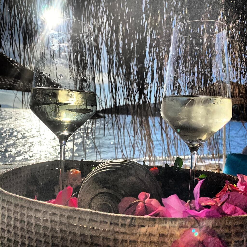 Two glasses of white wine on a table with a view of a body of water and a wooden structure in the background.