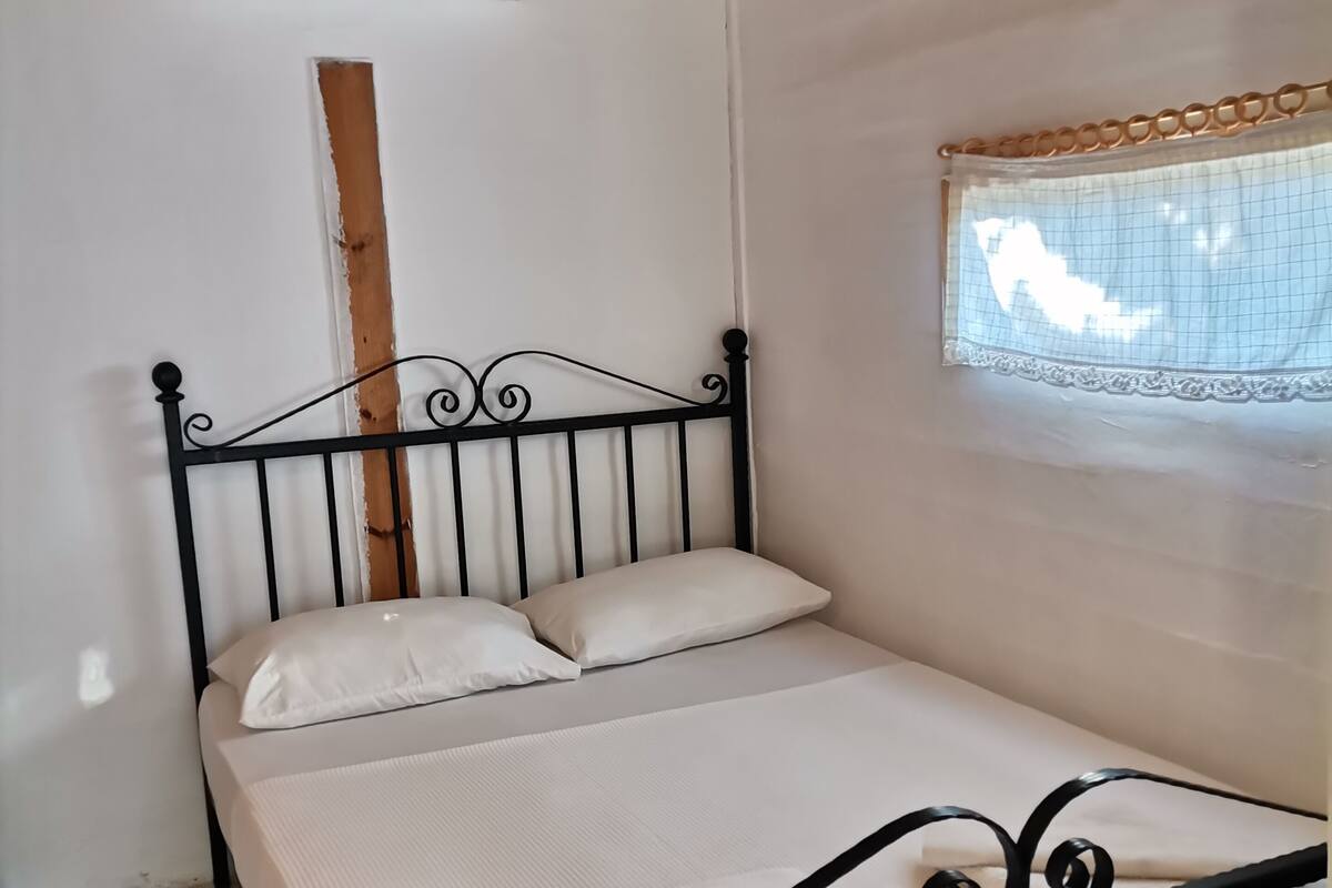 A single bed with a metal frame and white bedding, a wooden ceiling, and a window with a curtain.