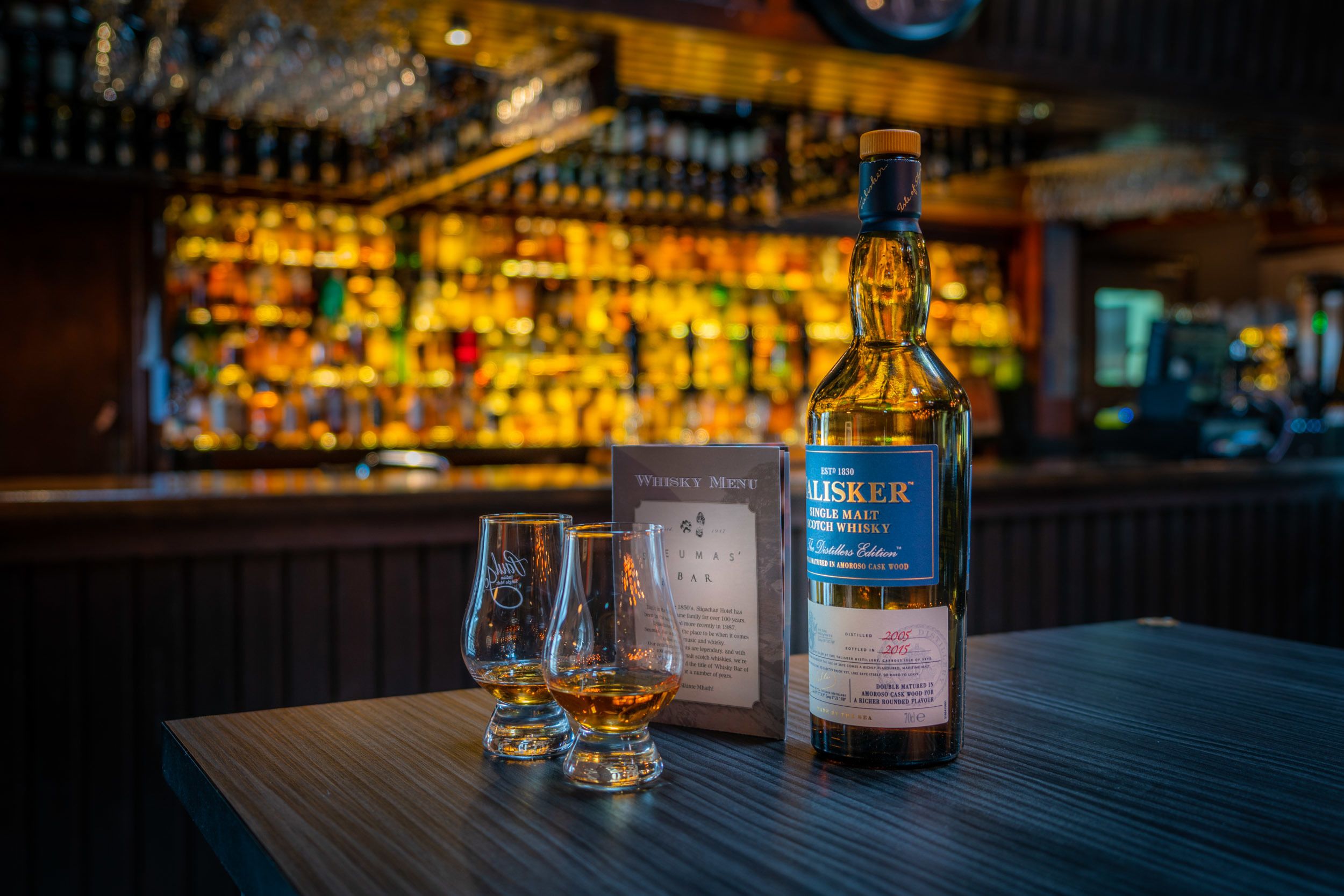A bottle of whisky and two glasses on a bar counter with a menu and a backdrop of a bar filled with bottles.