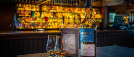 A bottle of whisky and two glasses on a bar counter with a menu and a backdrop of a bar filled with bottles.