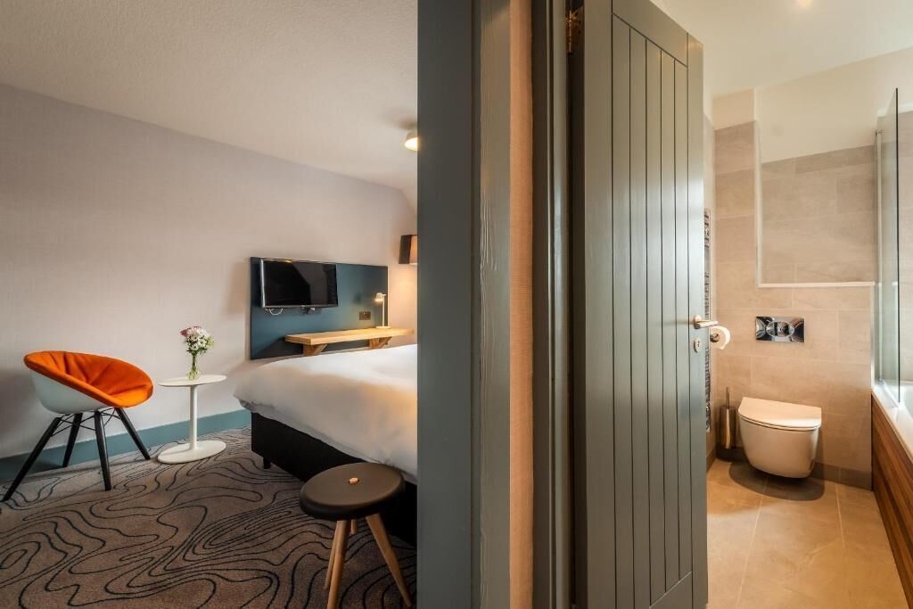 Une chambre d’hôtel comprenant un lit, une télévision, une petite table avec un vase, une chaise et une salle de bain avec des toilettes et une douche.