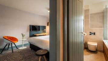 Une chambre d’hôtel comprenant un lit, une télévision, une petite table avec un vase, une chaise et une salle de bain avec des toilettes et une douche.
