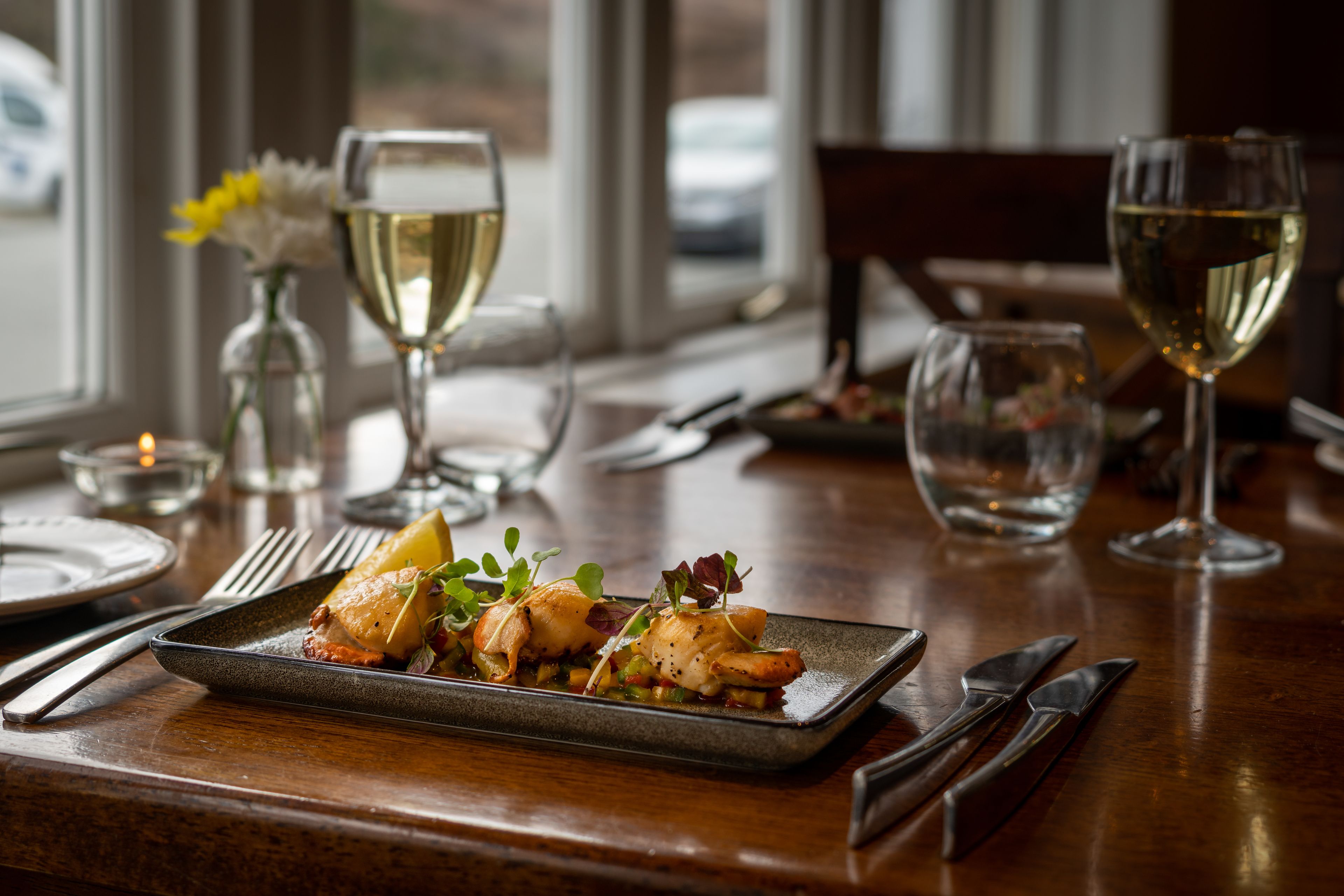 A dining table with a plated dish, wine glasses, cutlery, and a vase with flowers.