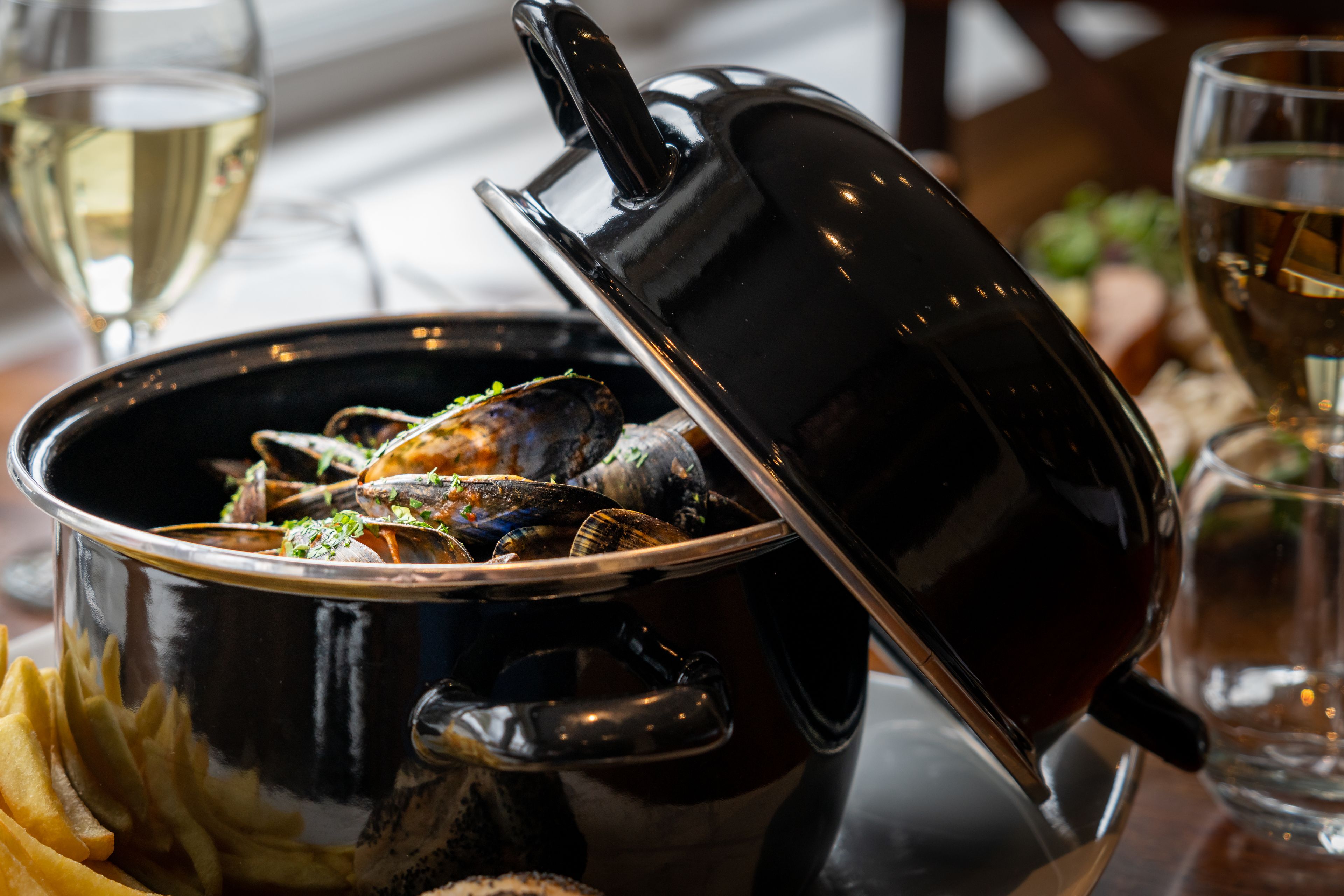 Une cocotte noire avec un couvercle, contenant des moules, accompagnée d’un verre de vin blanc et d’un verre de vin rouge.
