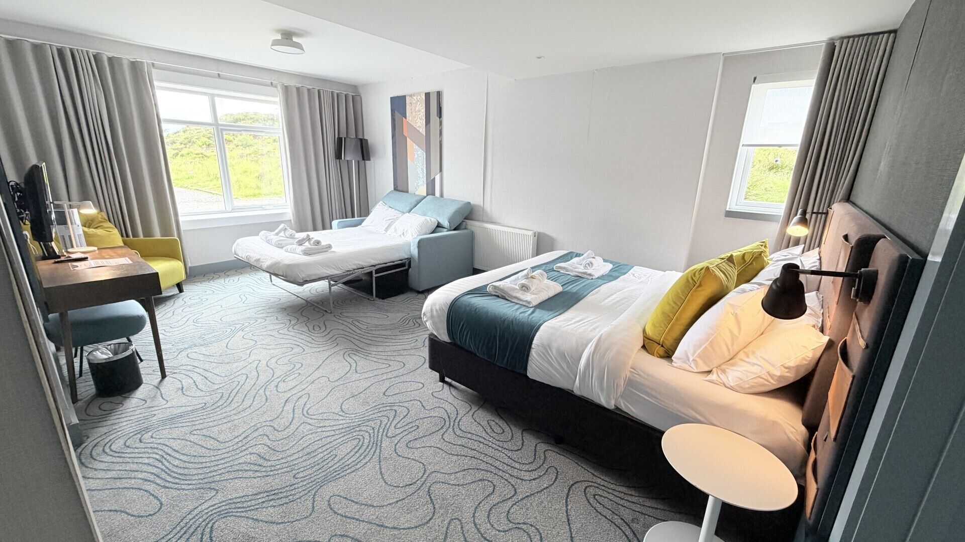 A modern hotel room with a bed, a desk, a chair, and a view of the outdoors.