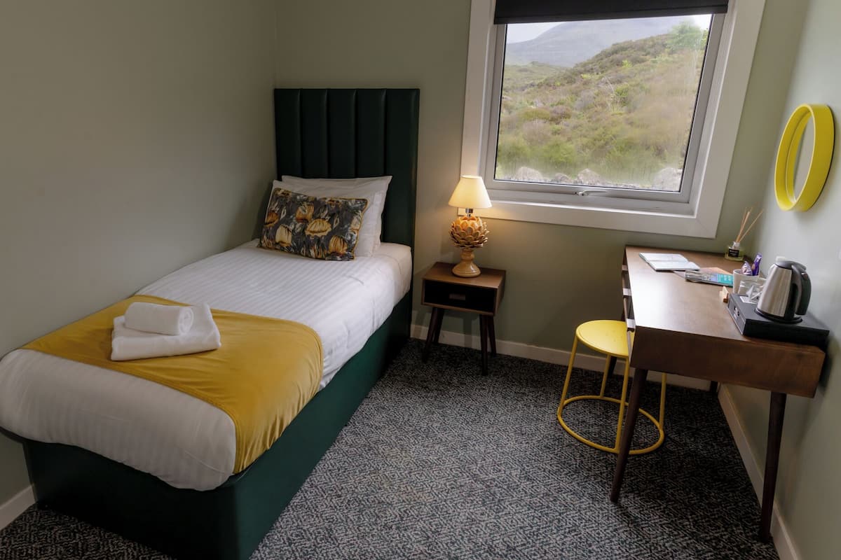 A neatly arranged bedroom with a bed, a bedside table with a lamp, a desk with a chair, and a view of the outdoors through a window.