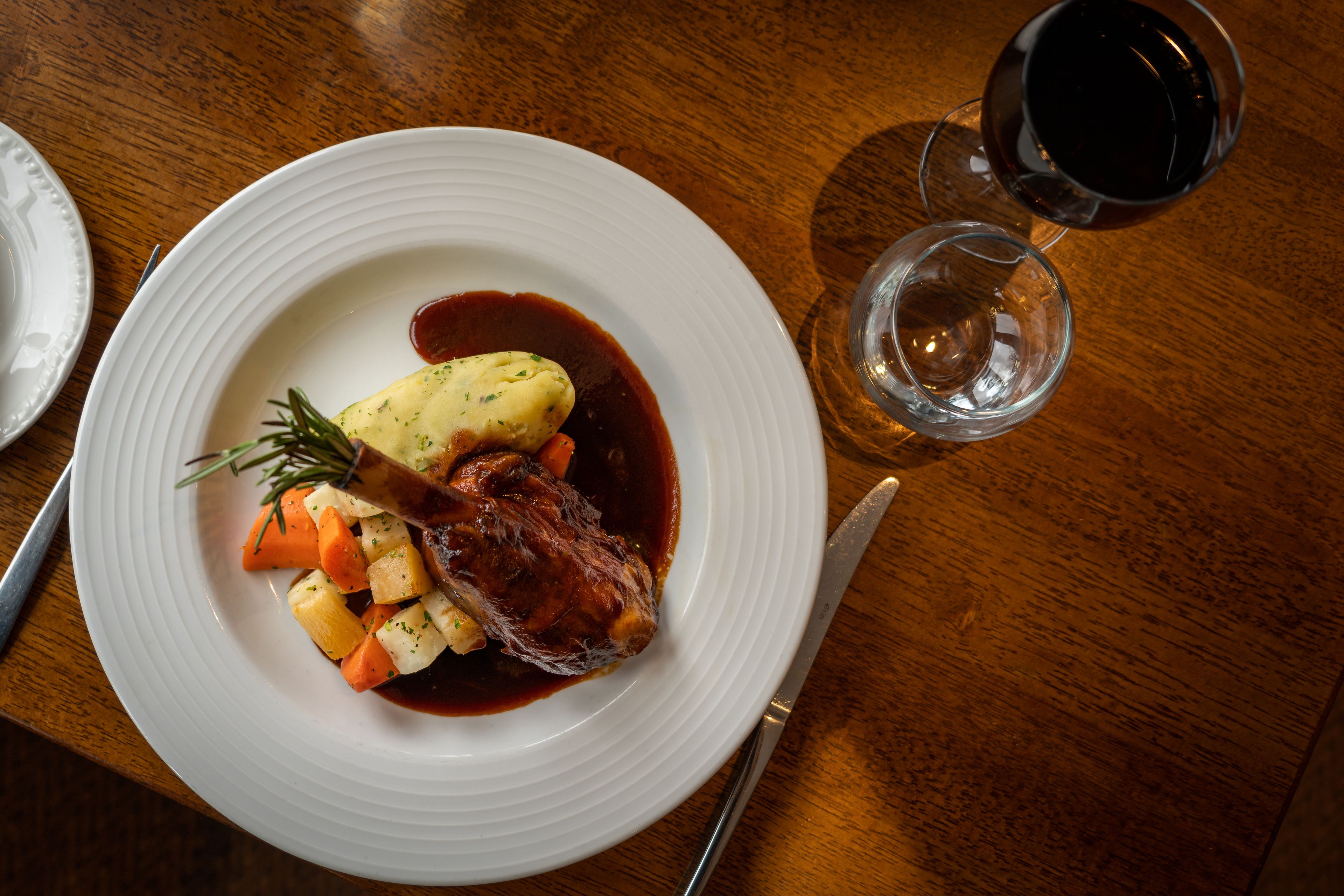 A plate of food with a meat dish, vegetables, and sauce, accompanied by a glass of red wine and a knife on a wooden table.