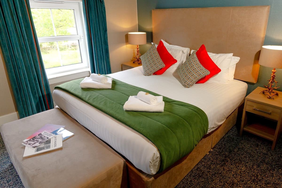 A neatly made bed with a green bedspread, white pillows, and red accent pillows. There are two bedside tables with lamps, a small table with a magazine, and a window with curtains.