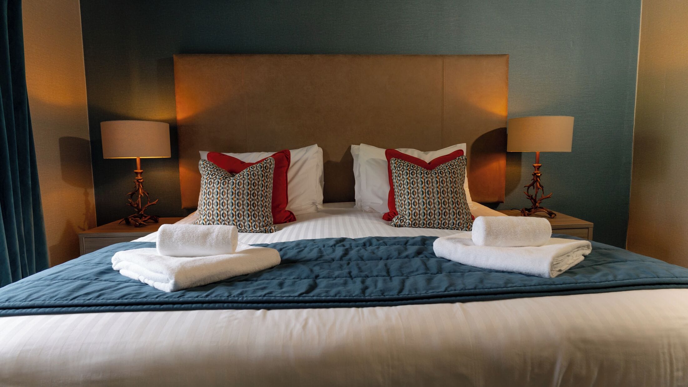 Un letto ben rifatto con una coperta blu, lenzuola bianche e due cuscini decorativi. Due abat-jour su ciascun lato del letto.