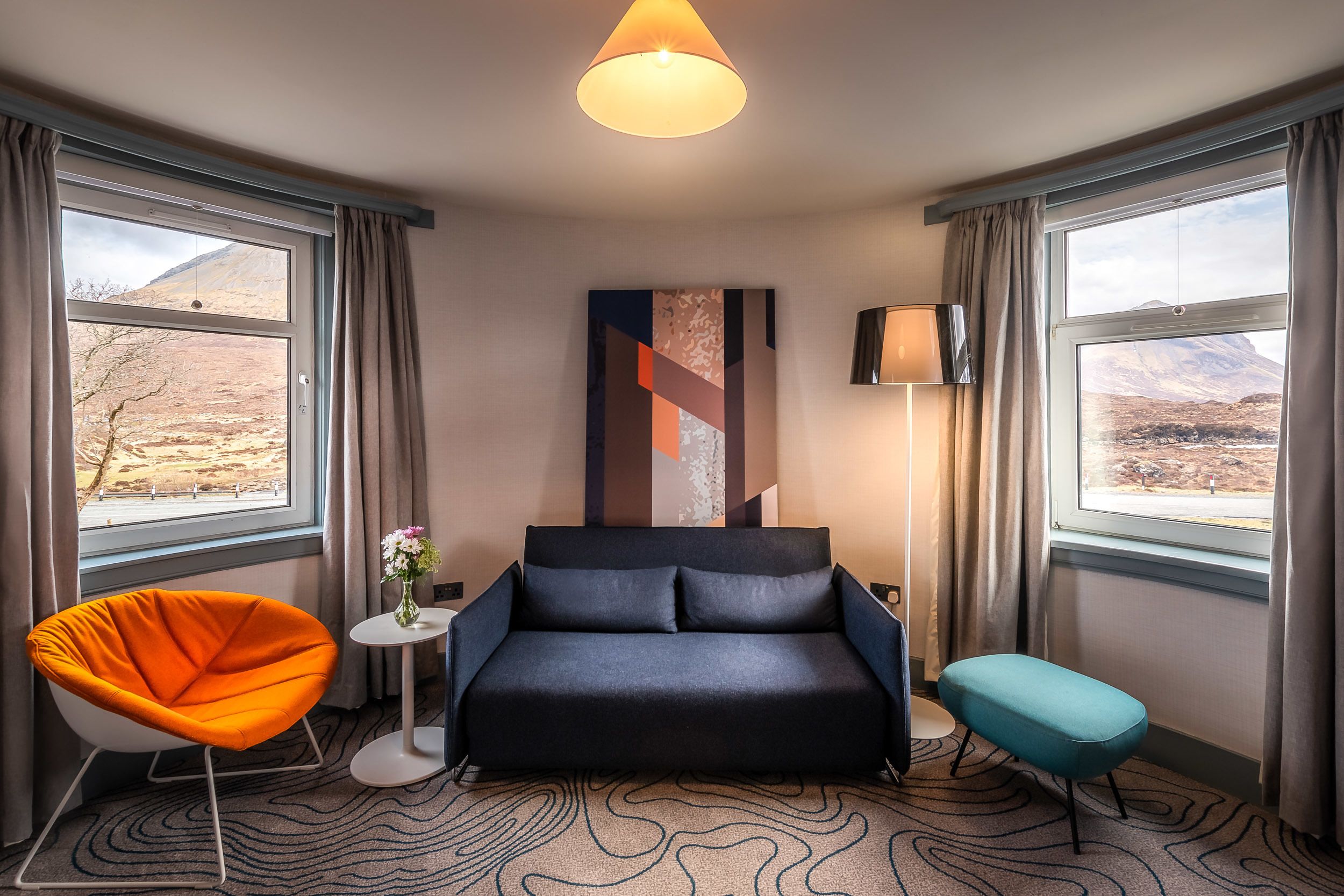 A modern hotel room with a dark blue sofa, an orange chair, a small round table with a vase of flowers, and a view of a mountainous landscape through large windows.