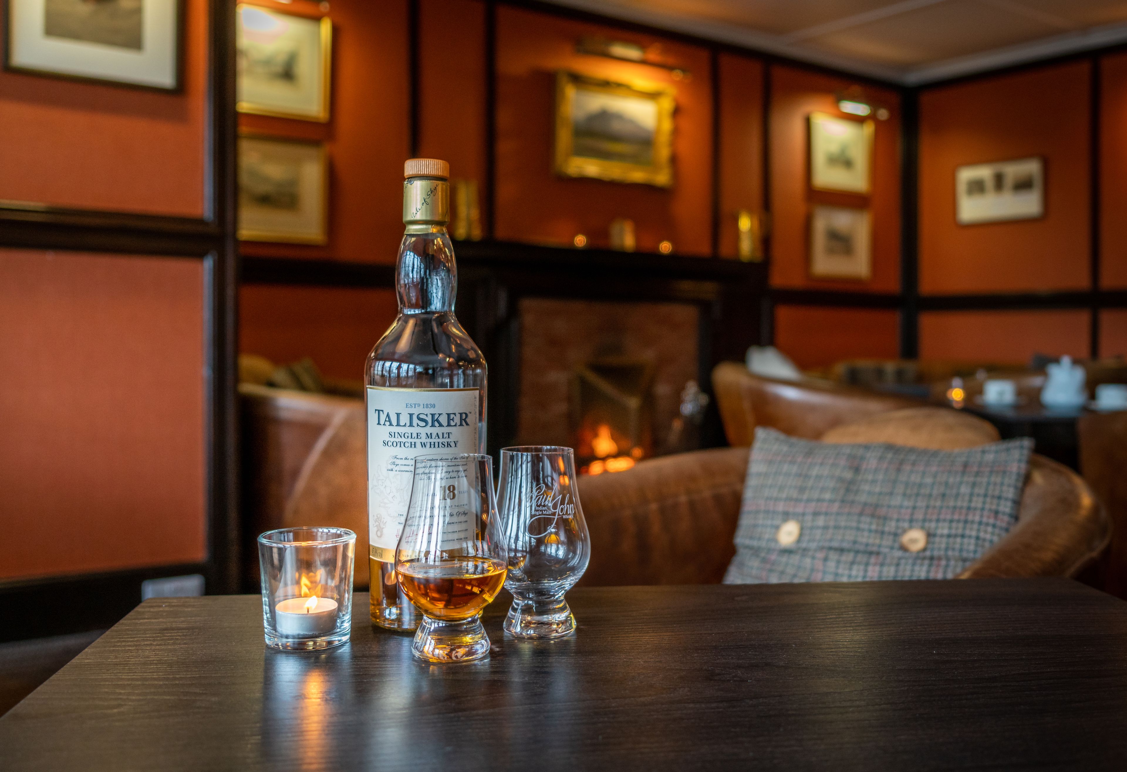 Una botella de whisky Talisker de malta única, dos vasos para licor y una vela encendida sobre una mesa en una habitación cálidamente iluminada con cuadros en las paredes.