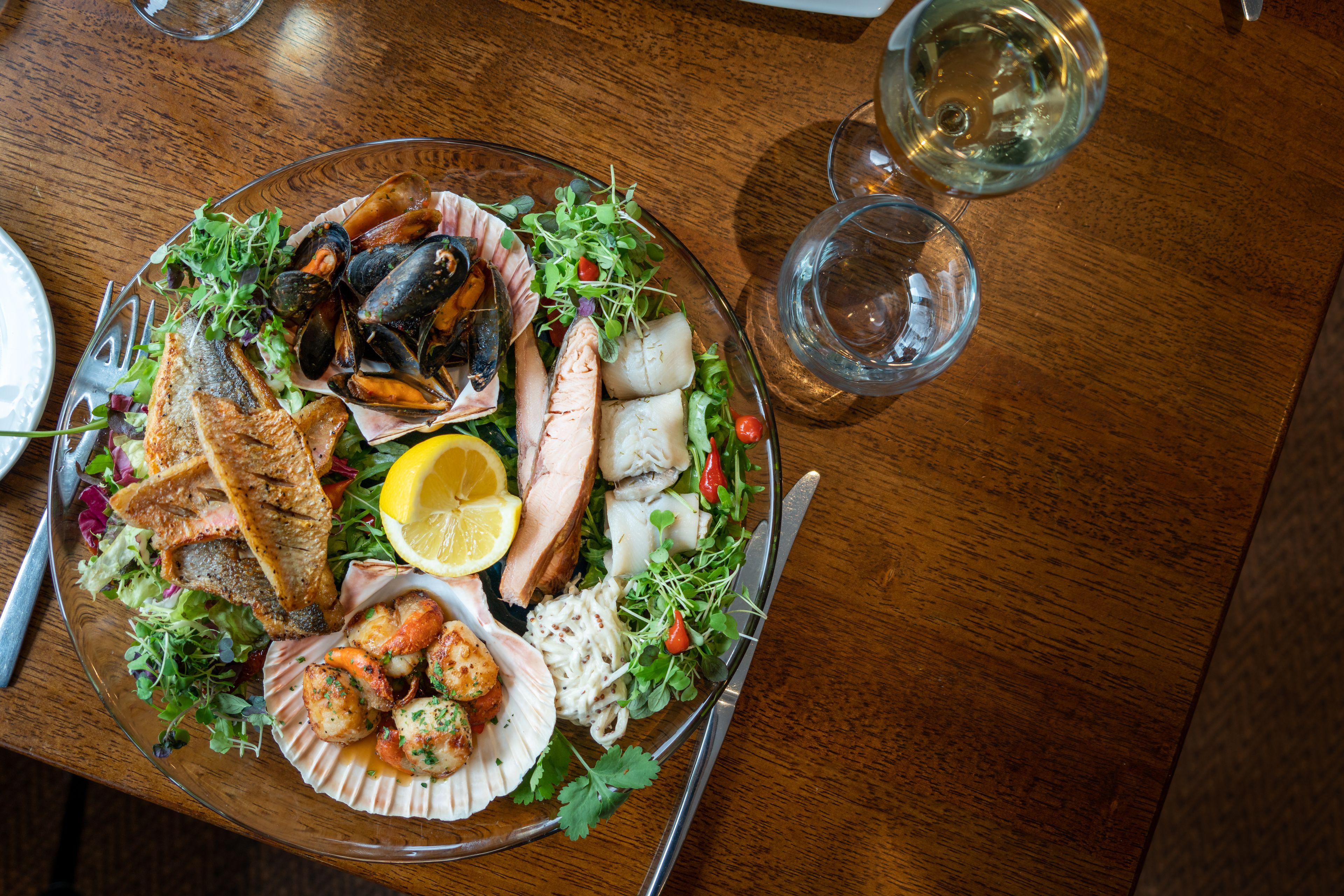 A seafood platter with various items like mussels, fish, and shrimp, accompanied by a glass of white wine and a glass of water on a wooden table.